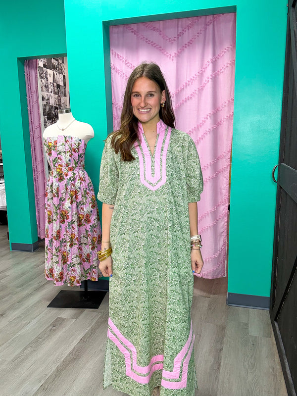 Woman in a green dress with pink trim standing next to a mannequin wearing a floral dress in a store setting.