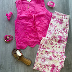 A styled flat-lay outfit on a gray wood floor featuring a bright pink sleeveless lace-trim top, cream pants with a pink floral print, pink high heels, a gold slide sandal, and pink accessories.