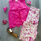 A styled flat-lay outfit on a gray wood floor featuring a bright pink sleeveless lace-trim top, cream pants with a pink floral print, pink high heels, a gold slide sandal, and pink accessories.