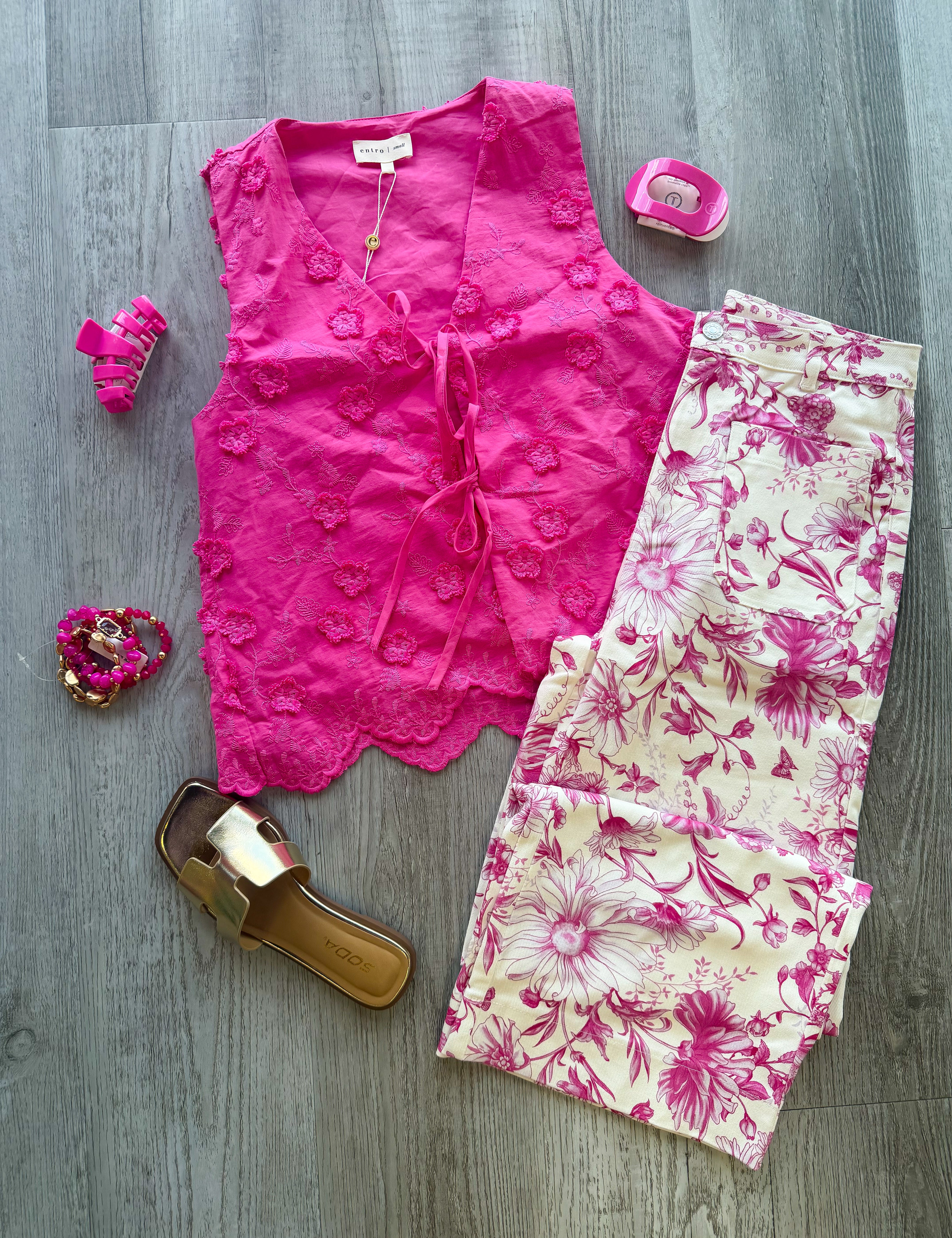 A styled flat-lay outfit on a gray wood floor featuring a bright pink sleeveless lace-trim top, cream pants with a pink floral print, pink high heels, a gold slide sandal, and pink accessories.