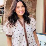 A woman wearing a cream-colored short-sleeve blouse with brown polka dots and brown trim, paired with blue jeans, standing outdoors in front of a stone building.