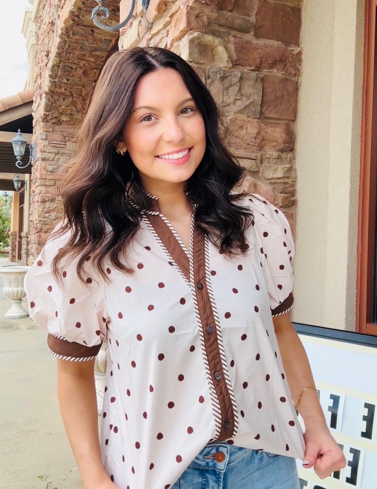 A woman wearing a cream-colored short-sleeve blouse with brown polka dots and brown trim, paired with blue jeans, standing outdoors in front of a stone building.