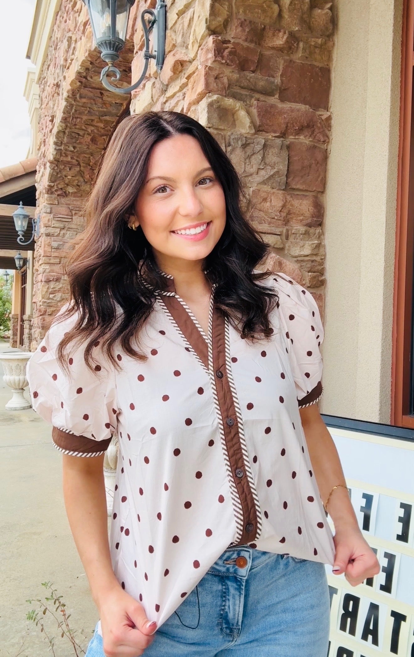 A woman wearing a cream-colored short-sleeve blouse with brown polka dots and brown trim, paired with blue jeans, standing outdoors in front of a stone building.