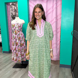 Woman in a green dress with pink trim standing next to a mannequin wearing a floral dress in a store setting.