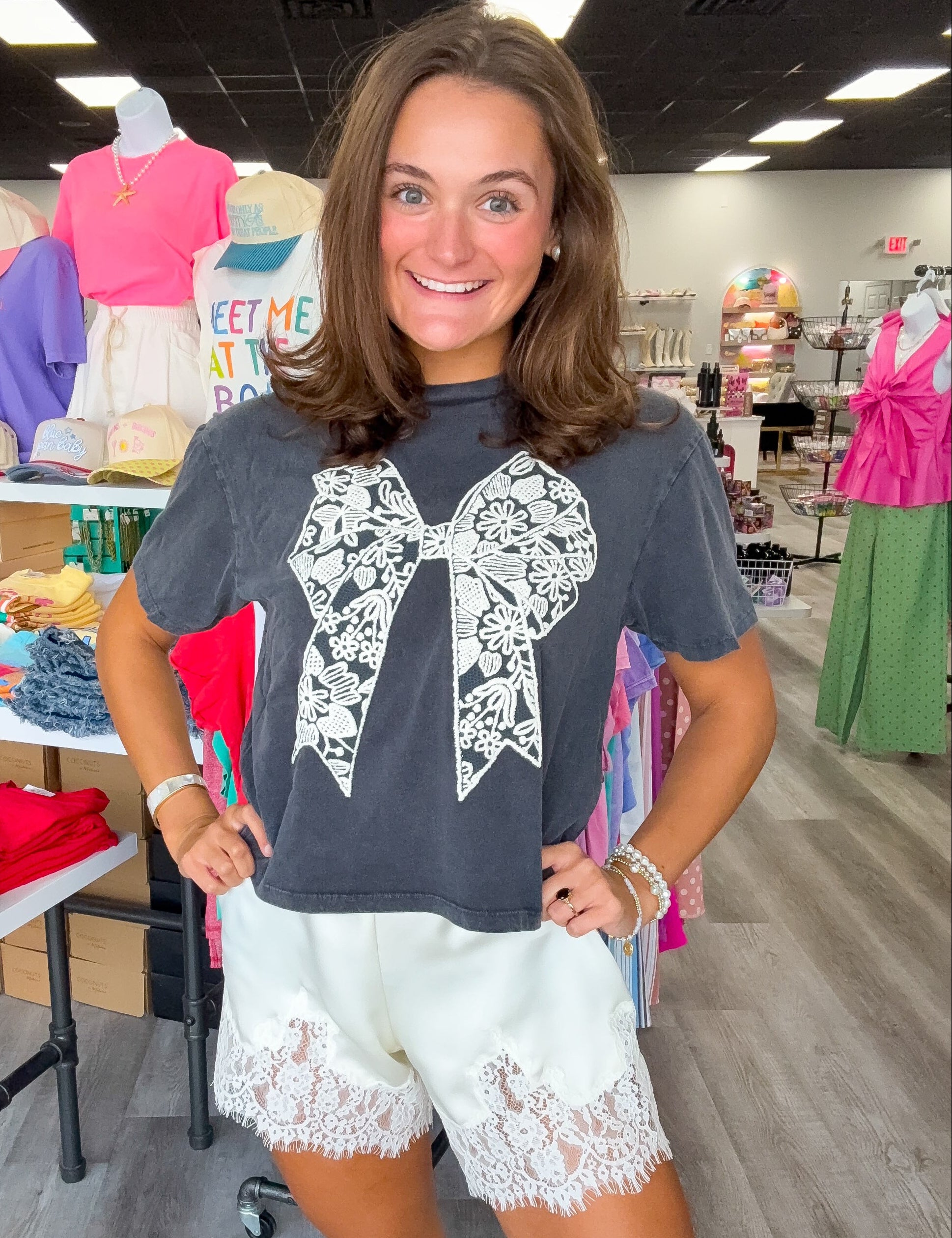 A woman modeling a dark gray t-shirt with a large white lace bow graphic on the front, paired with white lace-trimmed shorts, inside a retail store.