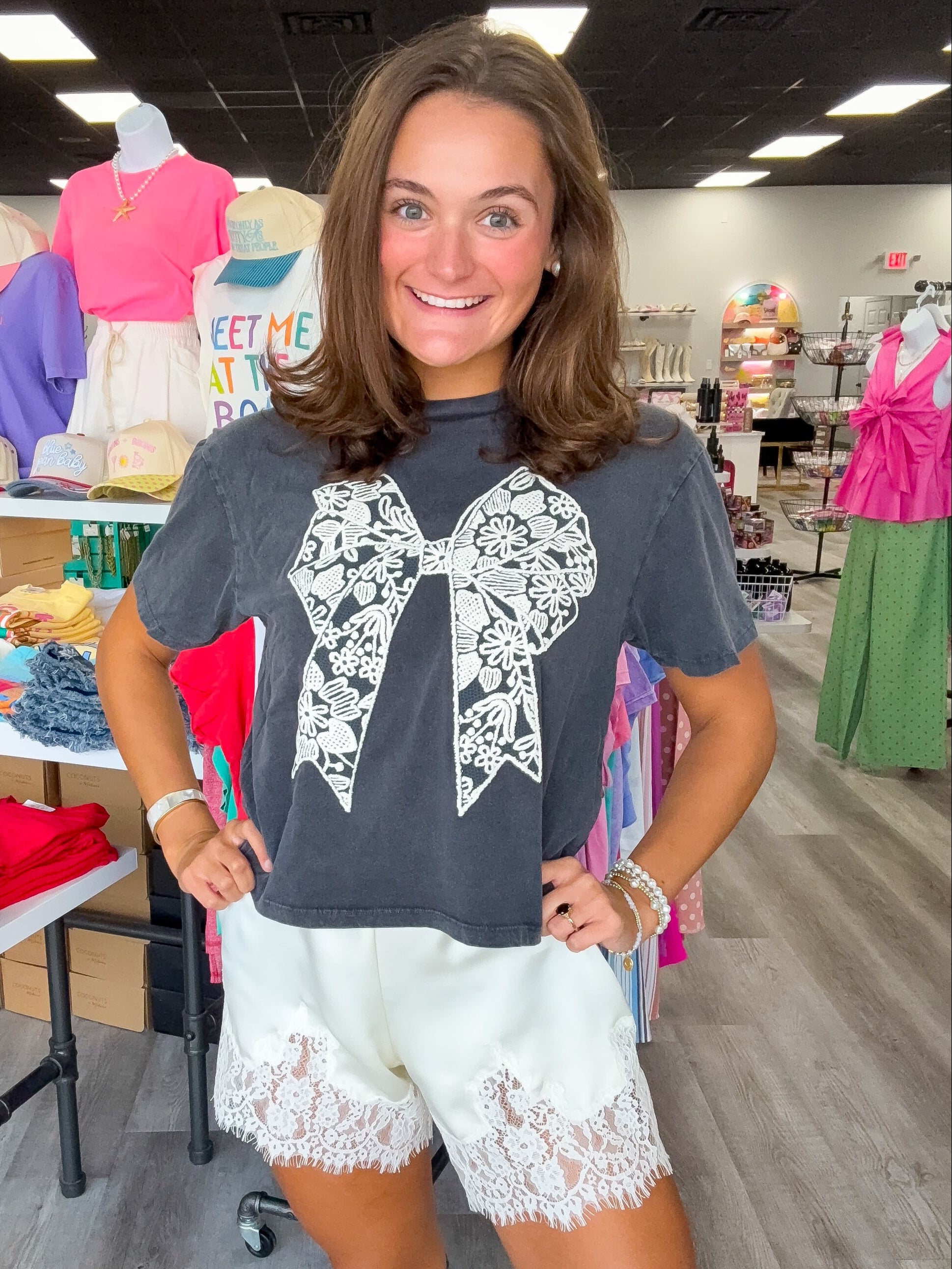 A woman modeling a dark gray t-shirt with a large white lace bow graphic on the front, paired with white lace-trimmed shorts, inside a retail store.