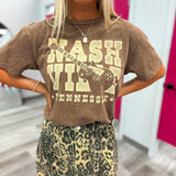 A woman wearing a brown vintage-style graphic t-shirt with a distressed cream 'Nashville Tennessee' and guitar print, paired with a leopard-print mini skirt, in what appears to be a fitting room.