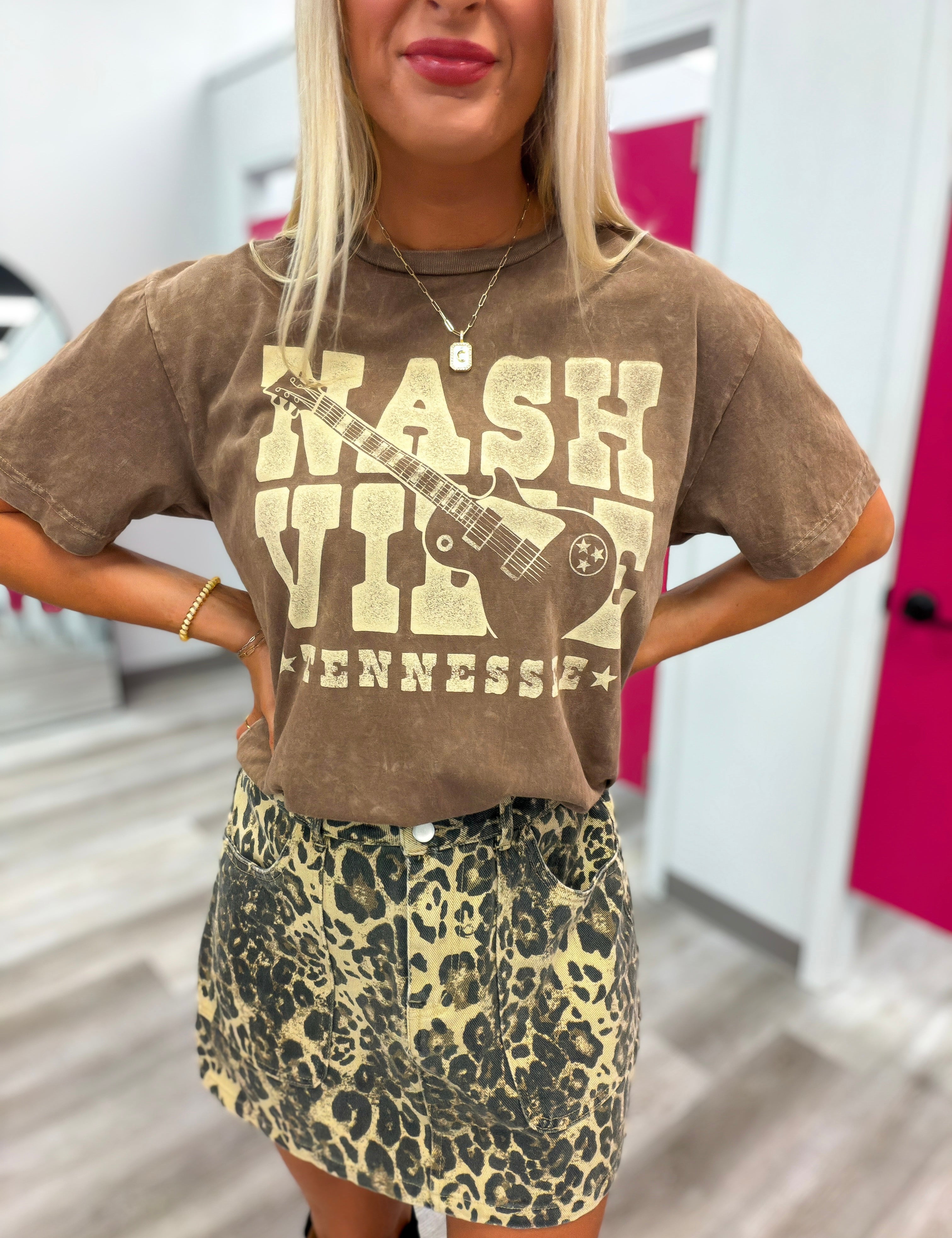 A woman wearing a brown vintage-style graphic t-shirt with a distressed cream 'Nashville Tennessee' and guitar print, paired with a leopard-print mini skirt, in what appears to be a fitting room.