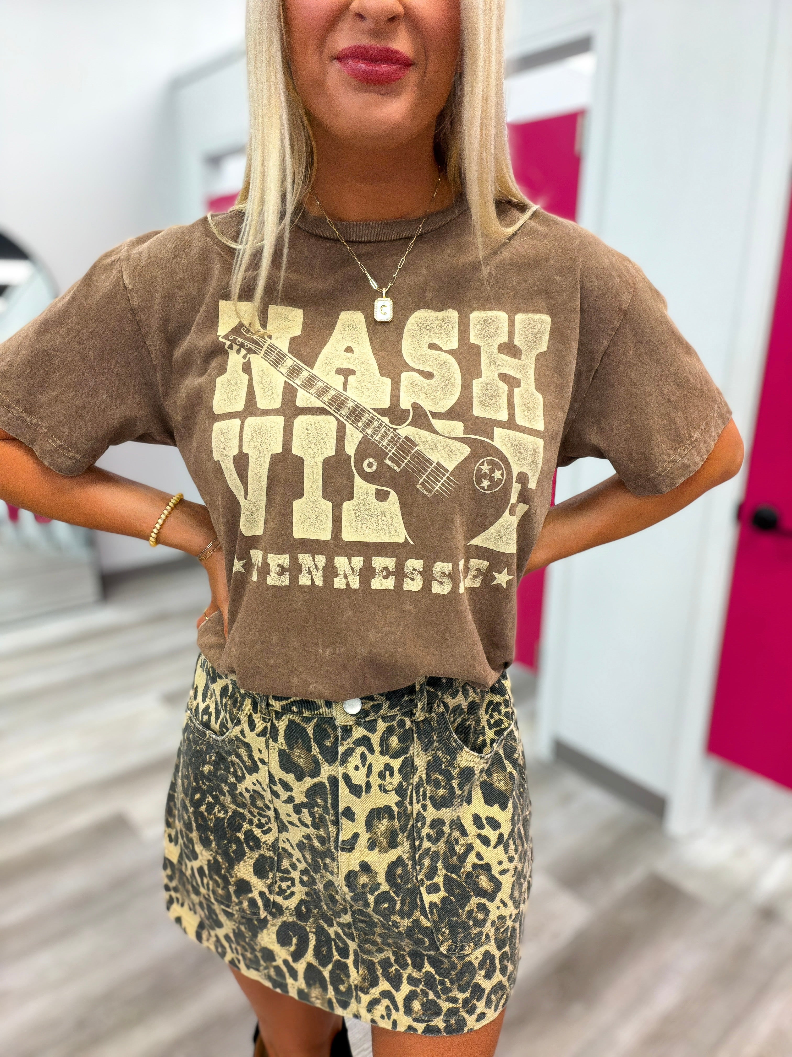 A woman wearing a brown vintage-style graphic t-shirt with a distressed cream 'Nashville Tennessee' and guitar print, paired with a leopard-print mini skirt, in what appears to be a fitting room.