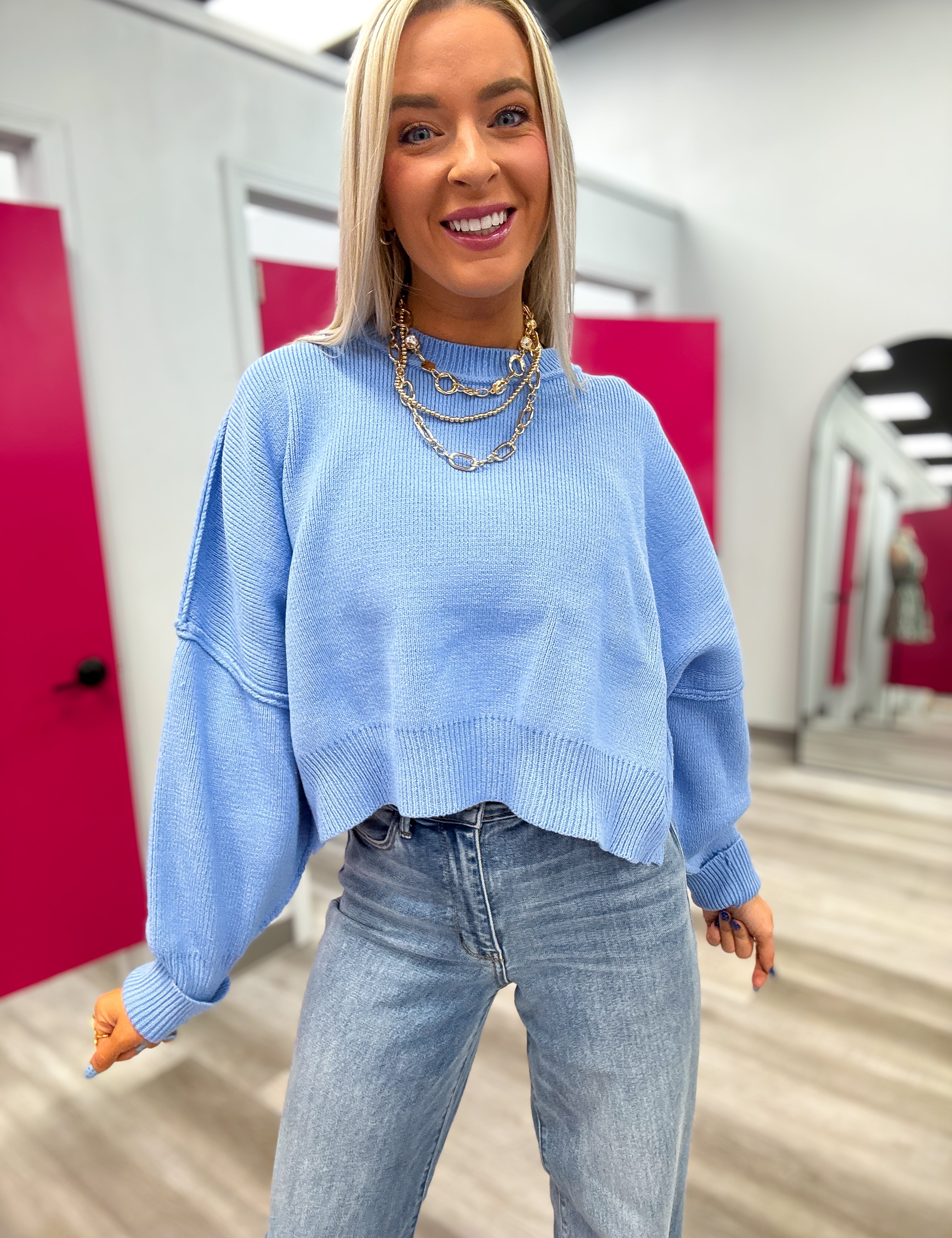 Woman wearing a light blue sweater and jeans in an indoor setting with pink walls.