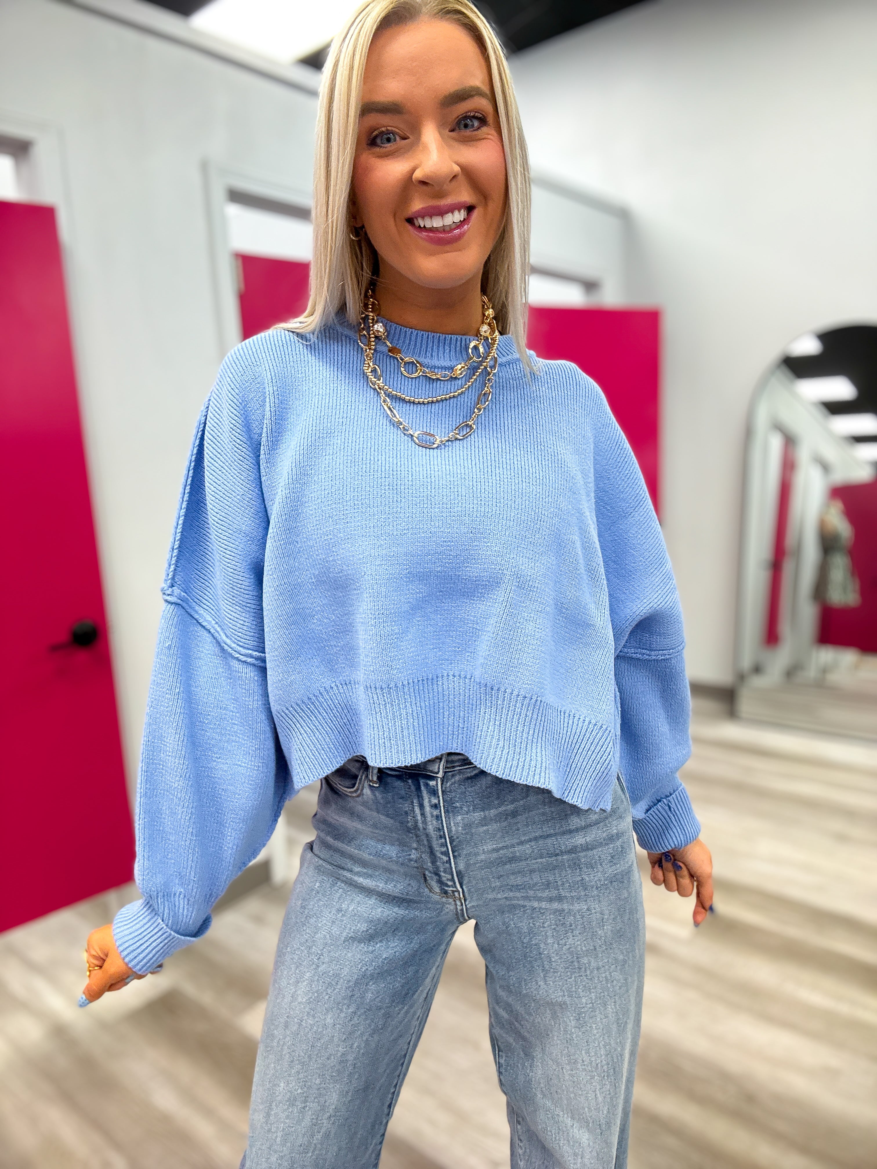 Woman wearing a light blue sweater and jeans in an indoor setting with pink walls.
