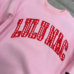 Pink sweatshirt with red 'LULU MAE' text on a gray surface