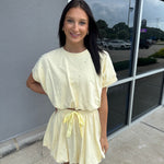 Woman in a yellow dress standing on a sidewalk with a reflective surface behind her.