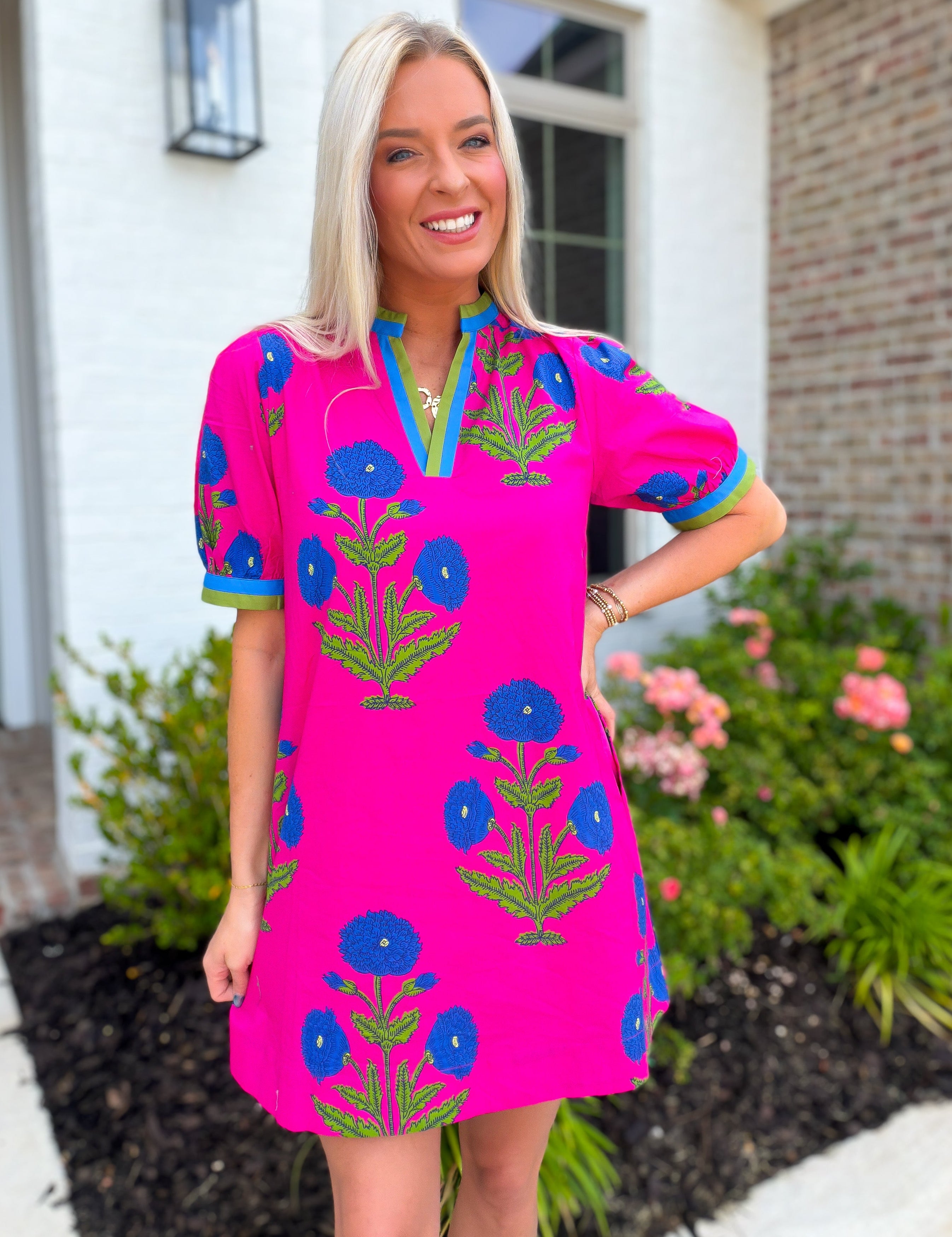 Woman wearing a bright pink dress with blue floral patterns standing in front of a house.