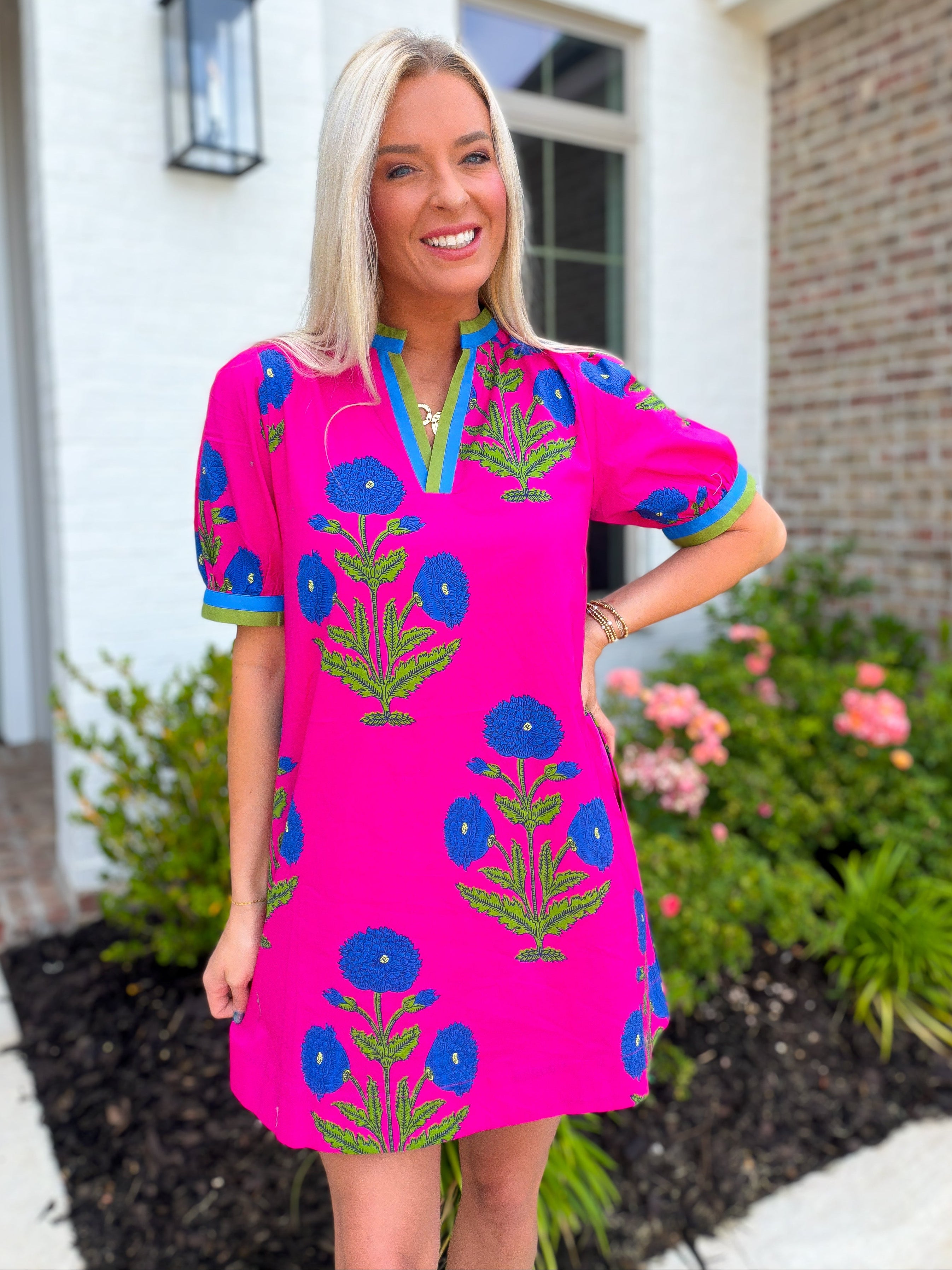 Woman wearing a bright pink dress with blue floral patterns standing in front of a house.